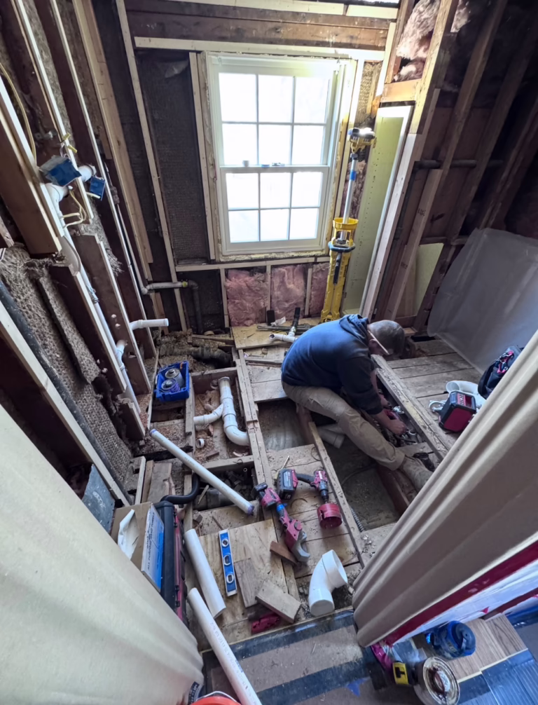Bathroom taken down to the studs and floor joists during demolition, with a plumber routing new drain lines through the joist space
