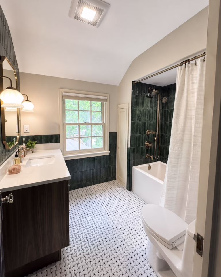 Finished remodeled bathroom with deep green vertical wall tile, white basketweave floor tile, a dark wood vanity, brass-toned fixtures, and a tub-shower