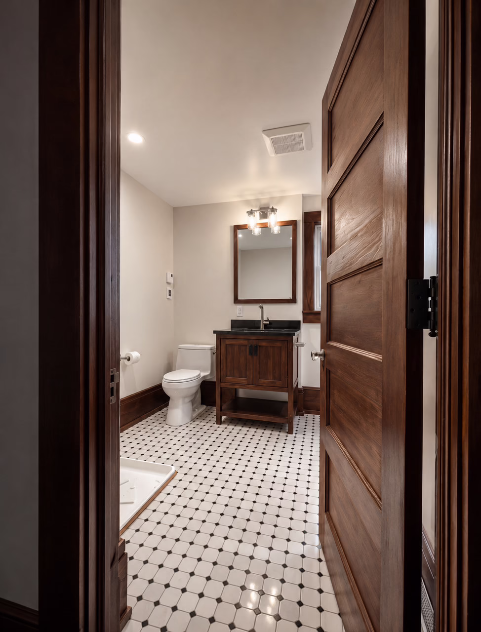 Finished bathroom with wood vanity, mirror, toilet, and black-and-white tile floor.