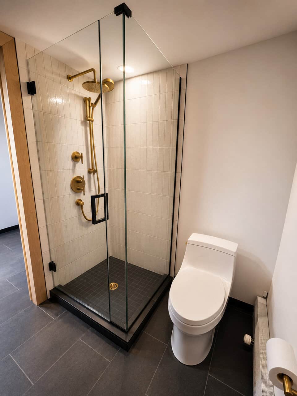 Glass shower with brass fixtures, beige tile, black base, white toilet, gray floor tile.
