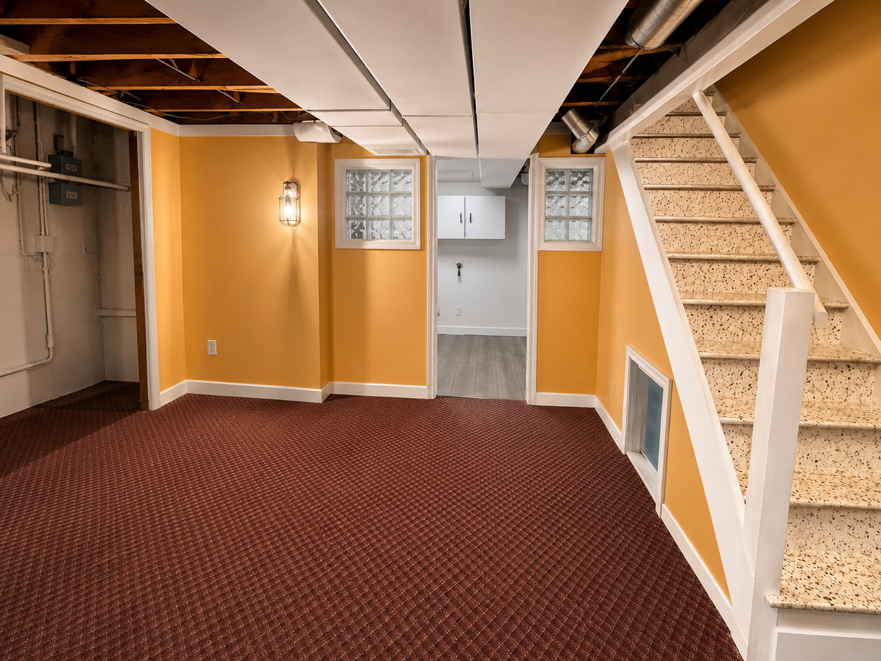 Dated basement with orange walls, red carpet, exposed ceiling, carpeted stairs.