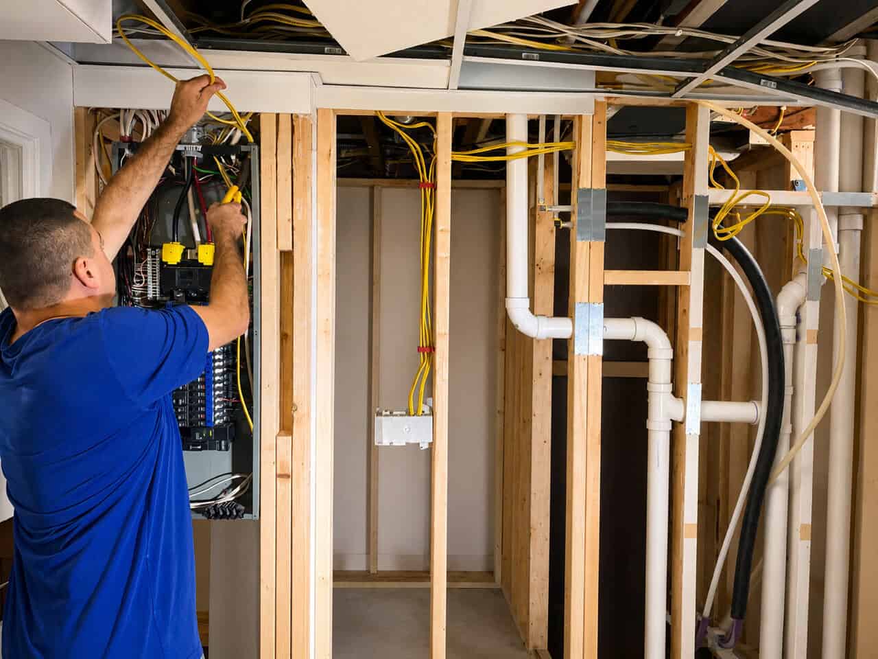 Electrician in blue shirt working on electrical panel near framed basement walls.
